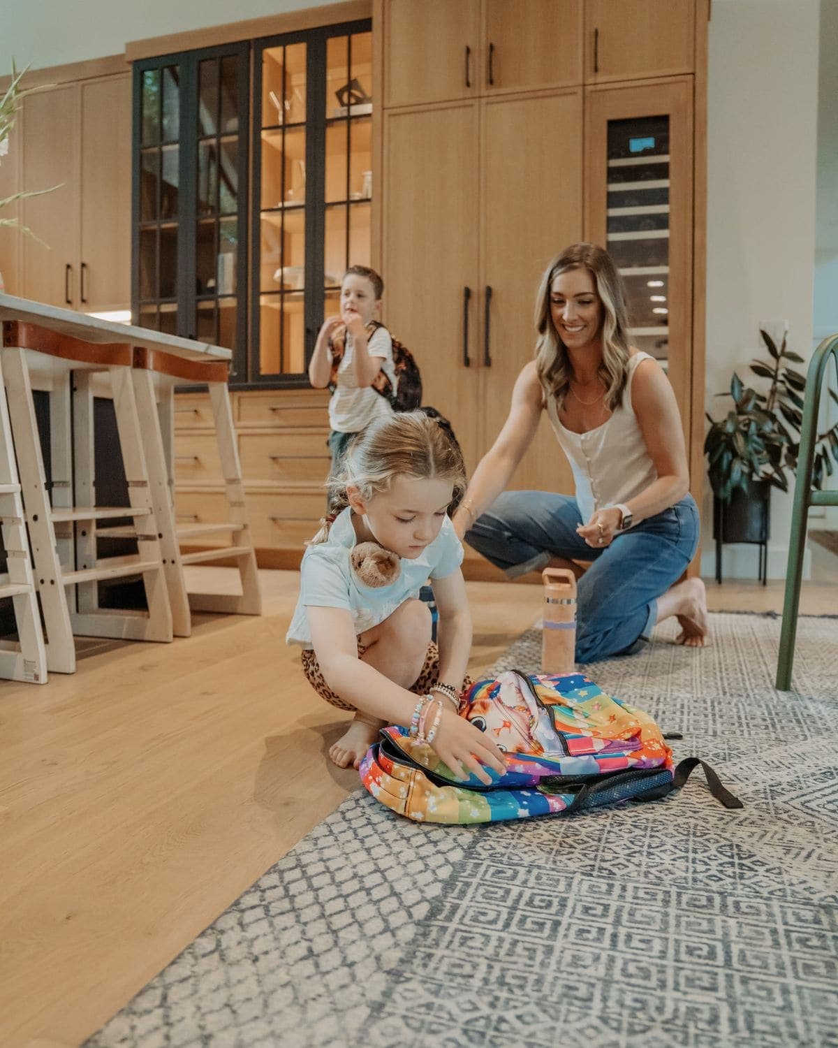 An adult helping two children with their school backpacks in the kitchen